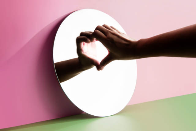 Hands forming a heart shape with their reflection in a round mirror, symbolising self-love and self-forgiveness