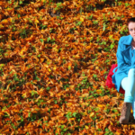 Woman in a blue coat sitting alone on a bench surrounded by a carpet of autumn leaves