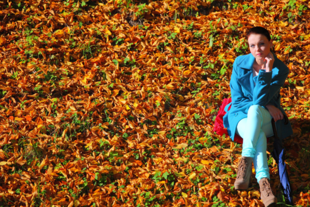Woman in a blue coat sitting alone on a bench surrounded by a carpet of autumn leaves